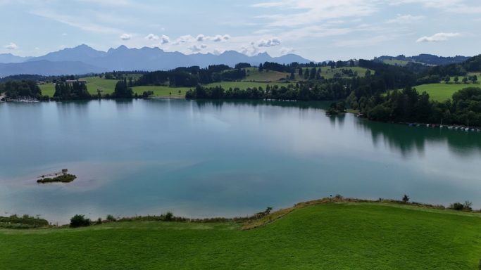 Ruhiger See umgeben von grünen Wiesen und Bergen im Hintergrund.